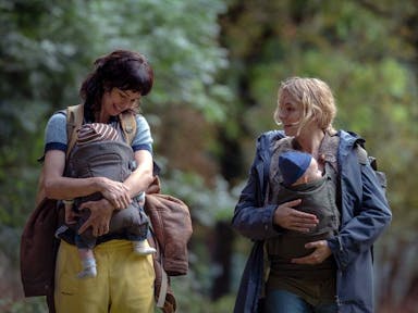 Two white women looking worn down, their babies strapped to their chests, carrying rucksacks as they walk through a woods