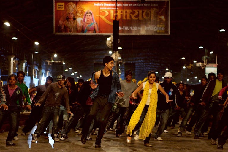 A young, slim Indian man and woman dancing energetically, with a  group of dancers lined behind them