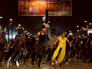 A young, slim Indian man and woman dancing energetically, with a group of dancers lined behind them