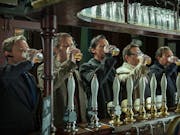 A line of 5 white men standing at a pubs bar all downing a pint of beer with their right hand