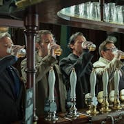 A line of 5 white men standing at a pubs bar all downing a pint of beer with their right hand