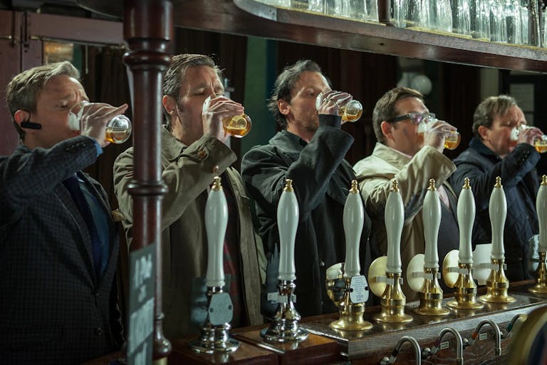 A line of 5 white men standing at a pubs bar all downing a pint of beer with their right hand