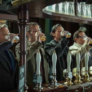 A line of 5 white men standing at a pubs bar all downing a pint of beer with their right hand