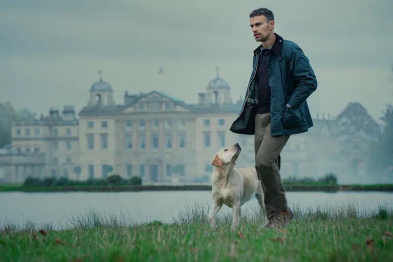 A white man in countryside attire, walks with a dog beside a lake with a grand estate behind him