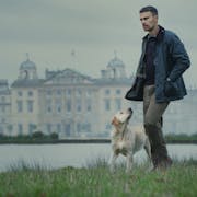 A white man in countryside attire, walks with a dog beside a lake with a grand estate behind him