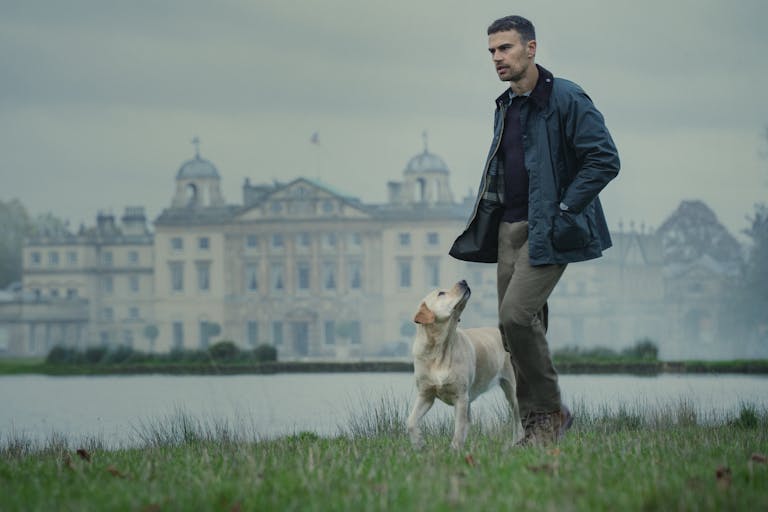 A white man in countryside attire, walks with a dog beside a lake with a grand estate behind him