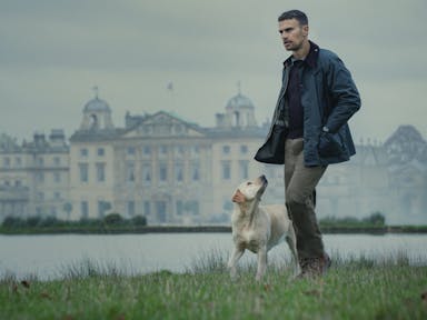 A white man in countryside attire, walks with a dog beside a lake with a grand estate behind him