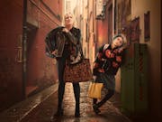 Woman and a child lean against an alleyway wall holding multiple bags