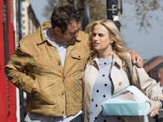 Man in beige jacket and blonde woman in white jacket walk down the street together, man has his arm around her shoulder.