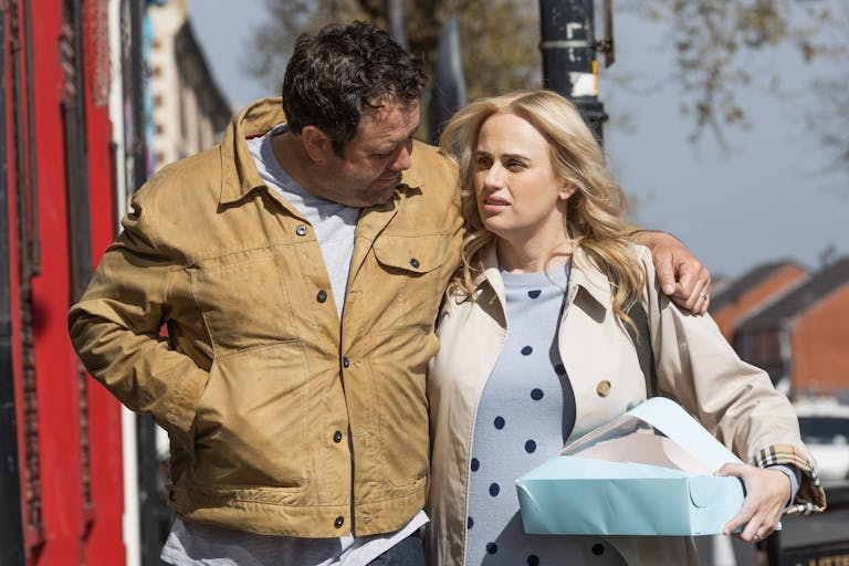 Man in beige jacket and blonde woman in white jacket walk down the street together, man has his arm around her shoulder. 