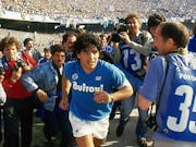 A young Argentinian man with black curly hair wearing a blue football shirt running through a crowd of photographers with a large stadium of supporters behind them