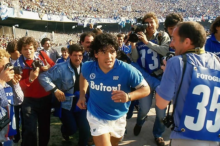 A young Argentinian man with black curly hair wearing a blue football shirt running through a crowd of photographers with a large stadium of supporters behind them