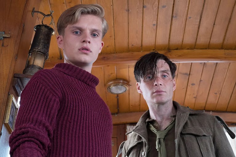 A young white man in a red turtleneck and a white man in ragged soldier's uniform in the inside of a small woodlined boat
