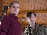 A young white man in a red turtleneck and a white man in ragged soldier's uniform in the inside of a small woodlined boat