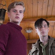 A young white man in a red turtleneck and a white man in ragged soldier's uniform in the inside of a small woodlined boat