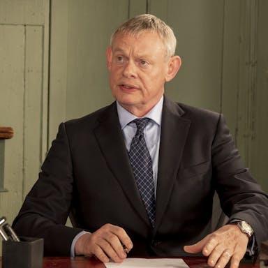 A white middle aged man with grey hair sitting at a desk in a suit with a quizzical facial expression 