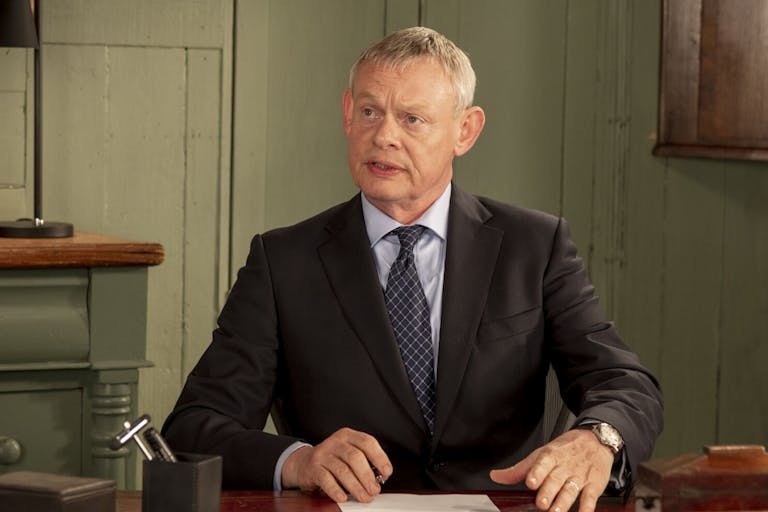 A white middle aged man with grey hair sitting at a desk in a suit with a quizzical facial expression