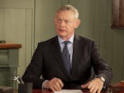 A white middle aged man with grey hair sitting at a desk in a suit with a quizzical facial expression