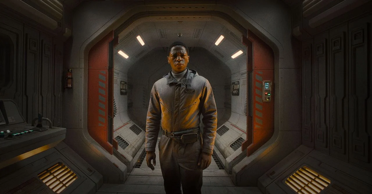 A young Black man in in brown overalls stands in a cinematic spaceship entrance looking shocked