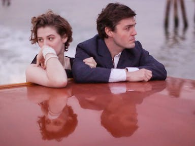 Young woman wearing a pearl braceley and a man in a suit stand arm in arm but looking in opposite directions on a boat. Their faces are reflected in the shiny red surface of the boat.