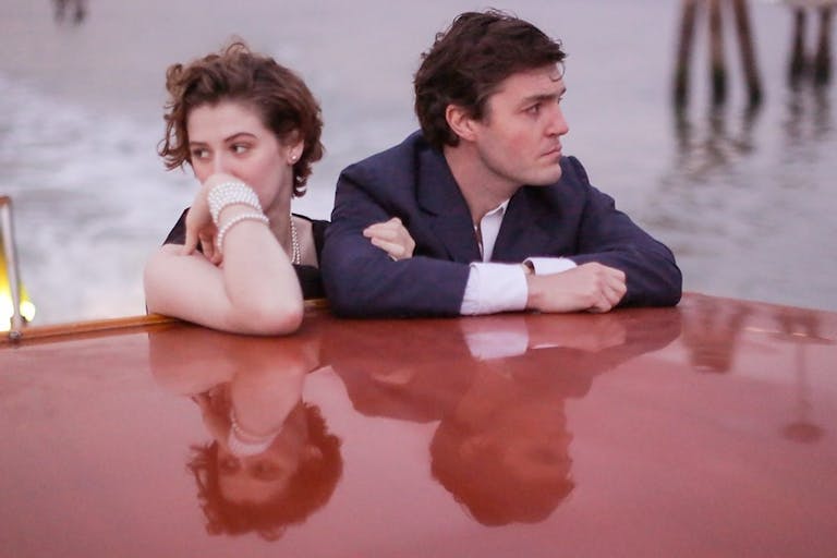 Young woman wearing a pearl braceley and a man in a suit stand arm in arm but looking in opposite directions on a boat. Their faces are reflected in the shiny red surface of the boat.