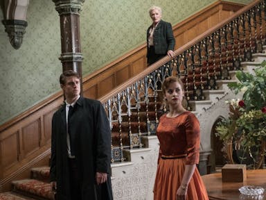 An older woman descending a grand staircase, in the foreground a man and a woman in smart clothes stand, all staring surprised 