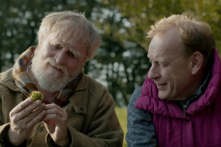 Two older white men sitting in a sunny field smiling looking a spiky acorn