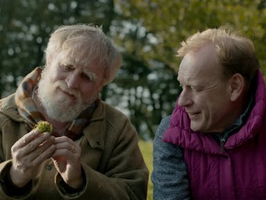 Two older white men sitting in a sunny field smiling looking a spiky acorn