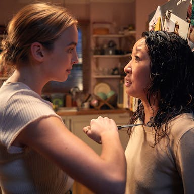 Blonde woman holds a woman with dark hair up against a fridge, knife in her hand