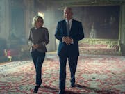 A white woman and a white man walk through a grand, royal room with a large oil painting behind them