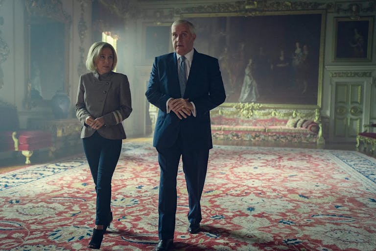 A white woman and a white man walk through a grand, royal room with a large oil painting behind them