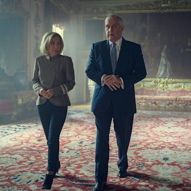 A white woman and a white man walk through a grand, royal room with a large oil painting behind them