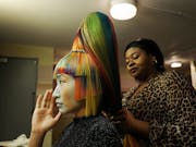 A woman with multi-coloured hair and sharp, angular chopped fringe and sides, with another woman behind her styling the hair into a tall bouffant