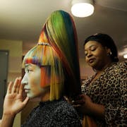 A woman with multi-coloured hair and sharp, angular chopped fringe and sides, with another woman behind her styling the hair into a tall bouffant