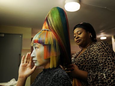 A woman with multi-coloured hair and sharp, angular chopped fringe and sides, with another woman behind her styling the hair into a tall bouffant