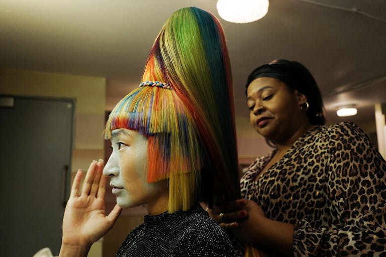 A woman with multi-coloured hair and sharp, angular chopped fringe and sides, with another woman behind her styling the hair into a tall bouffant