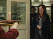 Middle aged woman with brown hair looks as if she will cry with joy as she walks into a room to someone holding a chocolate cake with a lit candle, she is followed by a smiling young man and there are balloons in the room.