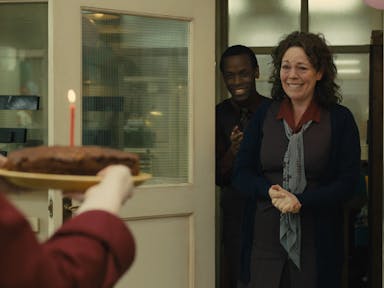 Middle aged woman with brown hair looks as if she will cry with joy as she walks into a room to someone holding a chocolate cake with a lit candle,  she is followed by a smiling young man and there are balloons in the room. 