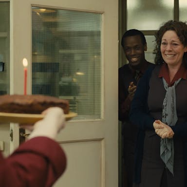 Middle aged woman with brown hair looks as if she will cry with joy as she walks into a room to someone holding a chocolate cake with a lit candle,  she is followed by a smiling young man and there are balloons in the room. 