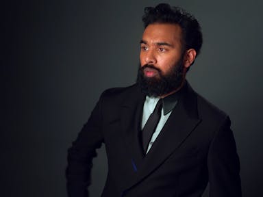 A brown man with dark hair and thick beard, looking suave in a black suit