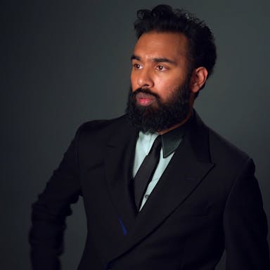 A brown man with dark hair and thick beard, looking suave in a black suit