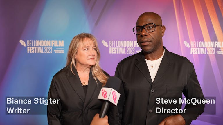 A white woman with blonde-brown hair, and a Black man with glasses talk into a microphone on the red carpet