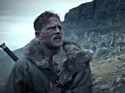 Blonde and bearded man wears a furry coat with a sword on his back, standing against a rugged and rocky landscape.