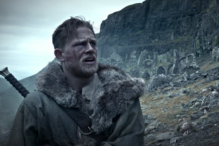 Blonde and bearded man wears a furry coat with a sword on his back, standing against a rugged and rocky landscape.