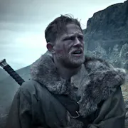 Blonde and bearded man wears a furry coat with a sword on his back, standing against a rugged and rocky landscape.