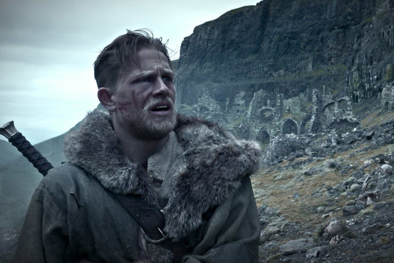 Blonde and bearded man wears a furry coat with a sword on his back, standing against a rugged and rocky landscape. 