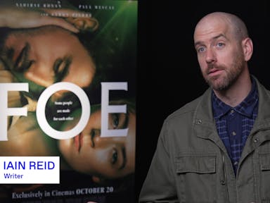A white man wearing a dark green jacket and blue shirt sits beside a film poster for Foe