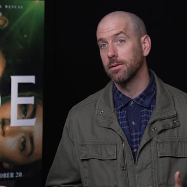 A white man wearing a dark green jacket and blue shirt sits beside a film poster for Foe