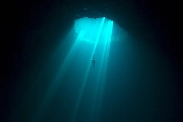 A diver in vast, dark body of water plunging through an opening looking lit by turquoise blue rays through the water