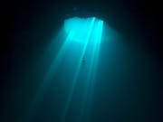 A diver in vast, dark body of water plunging through an opening looking lit by turquoise blue rays through the water
